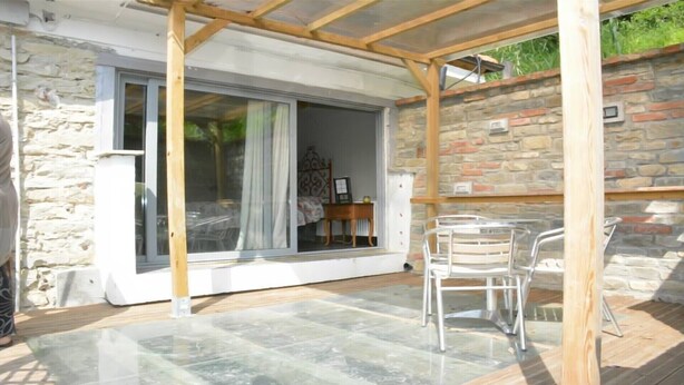 A covered patio area with a stone wall, a wooden pergola, and a glass door leading to a room with a bed and a table.