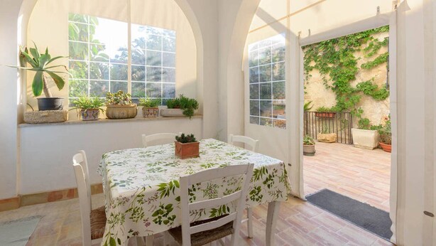 A room with a table and chairs, large arched windows, and potted plants.