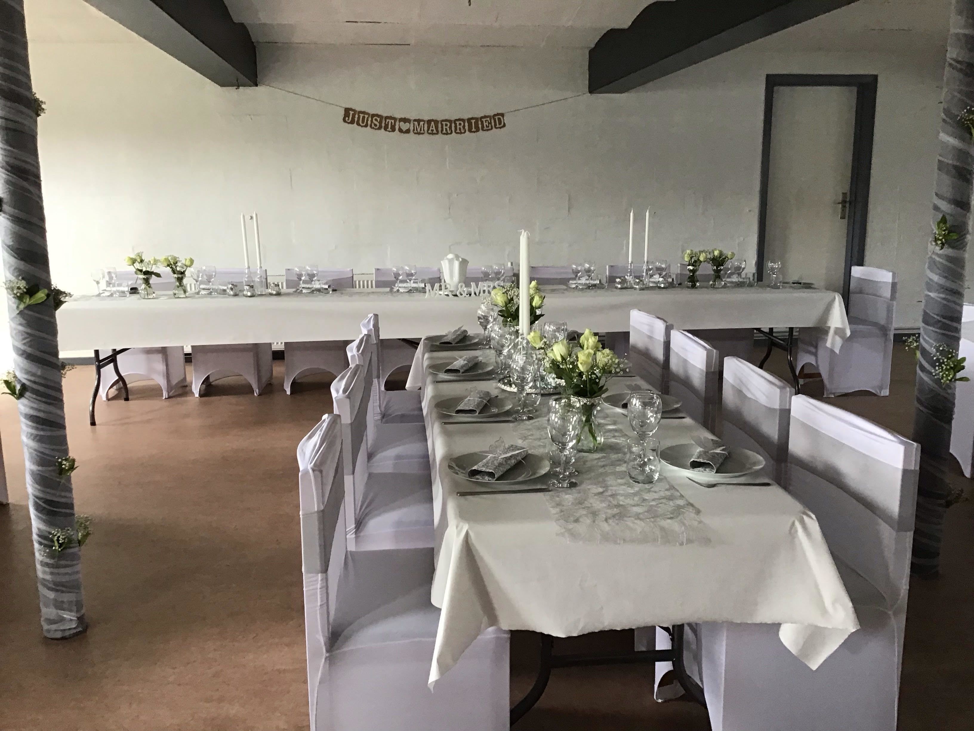 Un salón de banquetes con largas mesas dispuestas para una recepción de boda, con manteles blancos, centros de mesa florales y sillas blancas.