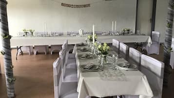 Un salón de banquetes con largas mesas dispuestas para una recepción de boda, con manteles blancos, centros de mesa florales y sillas blancas.