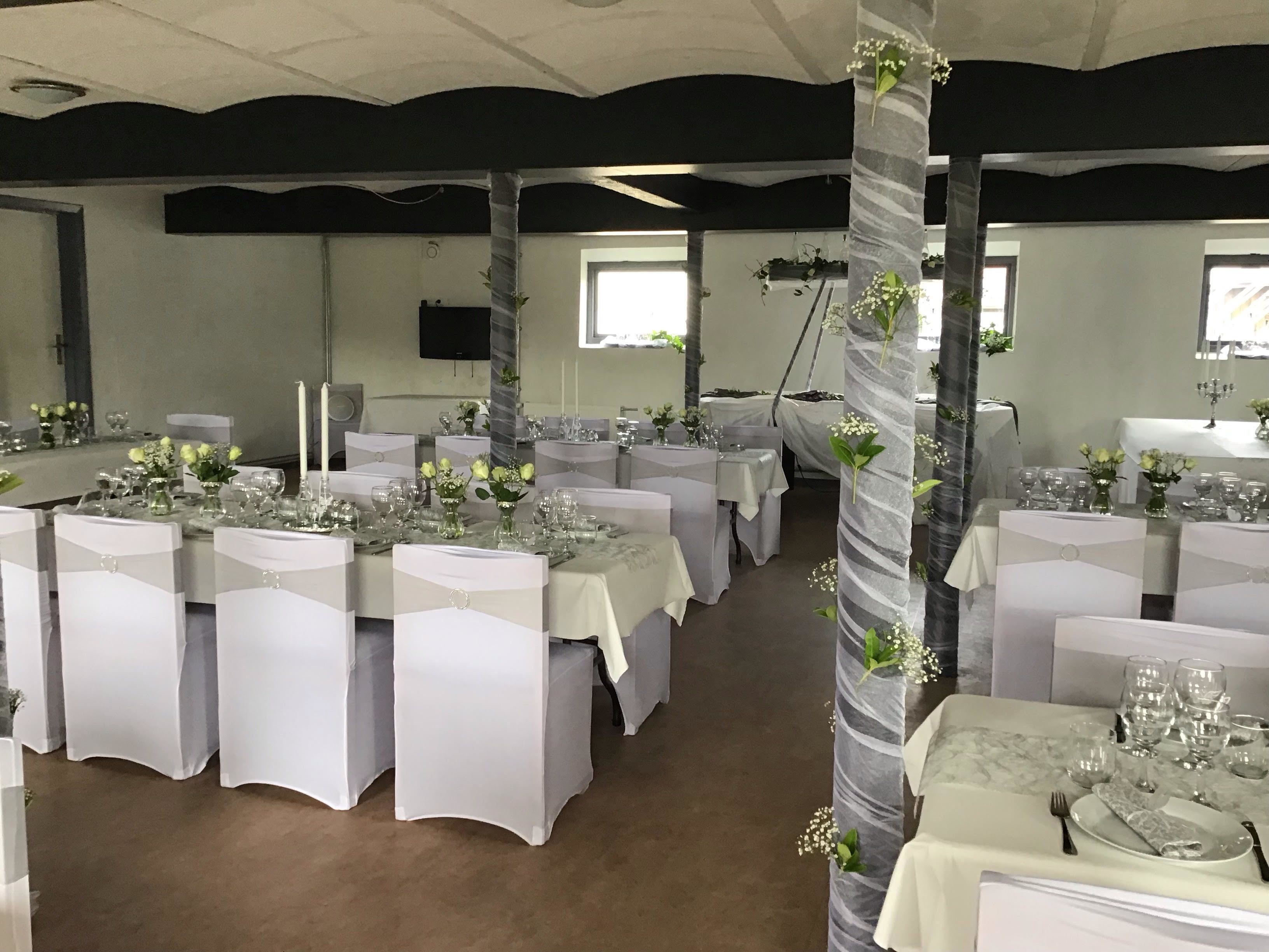 Salão de festas com várias mesas redondas dispostas com toalhas brancas, copos e arranjos florais.