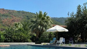 Una piscina cristalina con sillas de descanso y sombrillas, rodeada de exuberante vegetación y colinas al fondo.