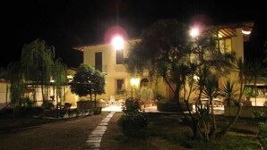 Vista nocturna de un edificio de dos plantas con un sendero pavimentado que conduce a la entrada, rodeado de jardines y árboles bien cuidados.
