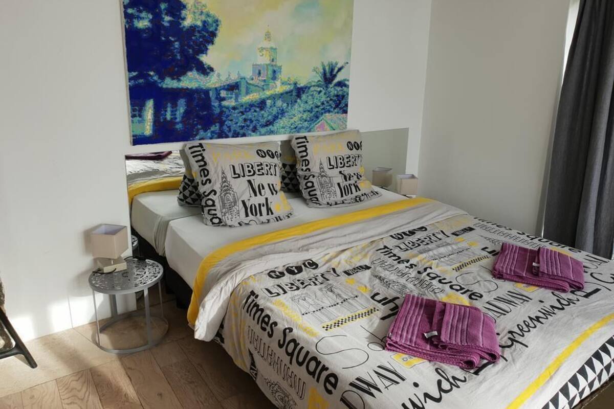 Ein Schlafzimmer mit einem Bett, das eine Bettdecke mit Times-Square-Motiv, Kissen mit den Aufdrucken 'Liberty' und 'New York' sowie einem kleinen runden Tisch mit einer Lampe zeigt. An der Wand hängt ein Kunstwerk in Blautönen.
