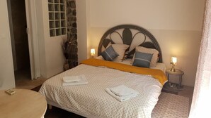 A neatly made bed with a patterned bedspread and pillows, a stone accent wall, a small bedside table with a lamp, and a door leading to another room. - K.N.C.1601 (La Croix-Valmer)