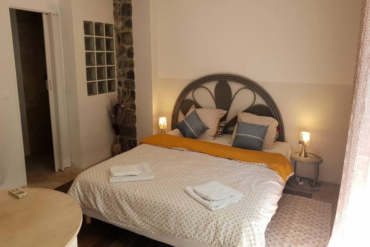 Ein ordentlich bezogenes Bett mit gemustertem Bettwäsche und Kissen, eine Wand mit Steinverkleidung, ein kleiner Nachttisch mit Lampe und eine Tür zu einem anderen Raum.