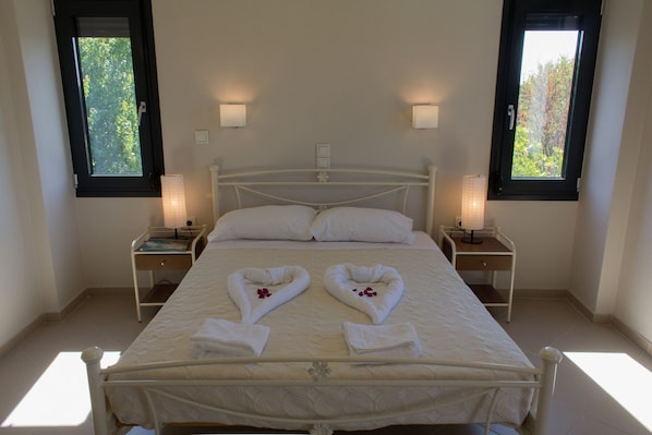 Una cama bien hecha con dos toallas con forma de corazón, dos lámparas de noche y dos ventanas con persianas.