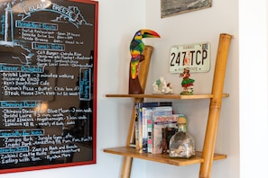 Un mur sur lequel est peint un tableau noir indiquant les endroits où prendre le petit-déjeuner, déjeuner et dîner, ainsi que les endroits où boire un verre le soir. On peut également apercevoir une étagère en bois avec des livres et une plaque d’immatriculation hawaïenne.