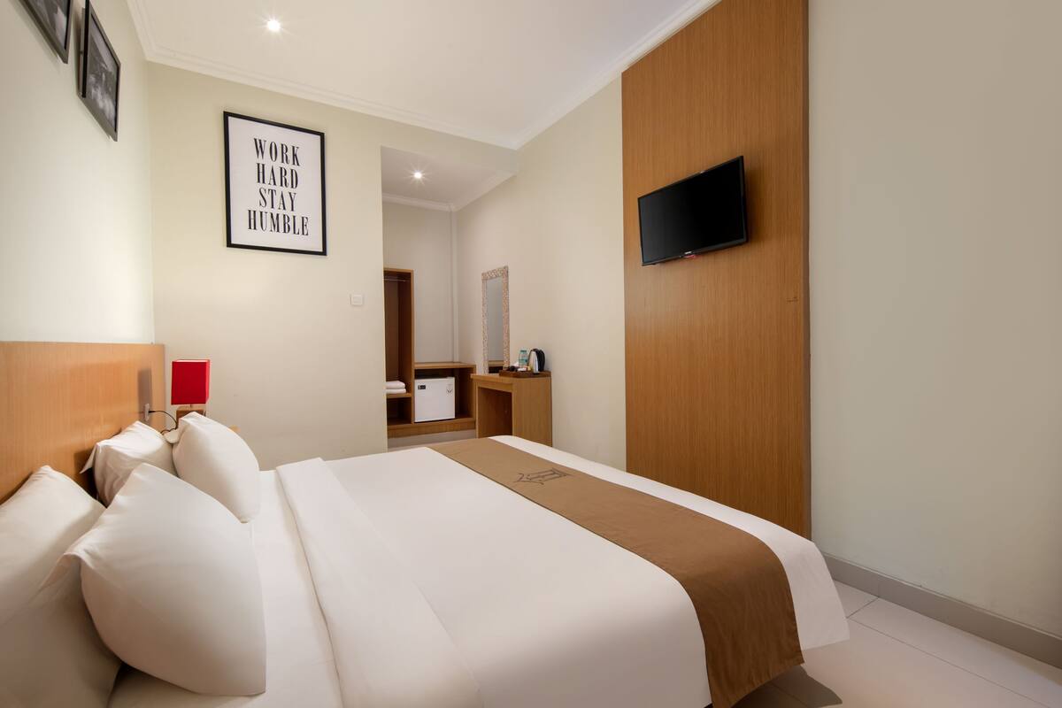 Une chambre d’hôtel avec un lit, un téléviseur fixé au mur, un miroir et une citation inspirante encadrée.