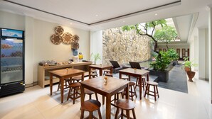 A modern dining area with wooden tables and chairs, a kitchen counter, and a stone wall with a tree. - Angkul Angkul Segara Beach Kuta (Kuta)