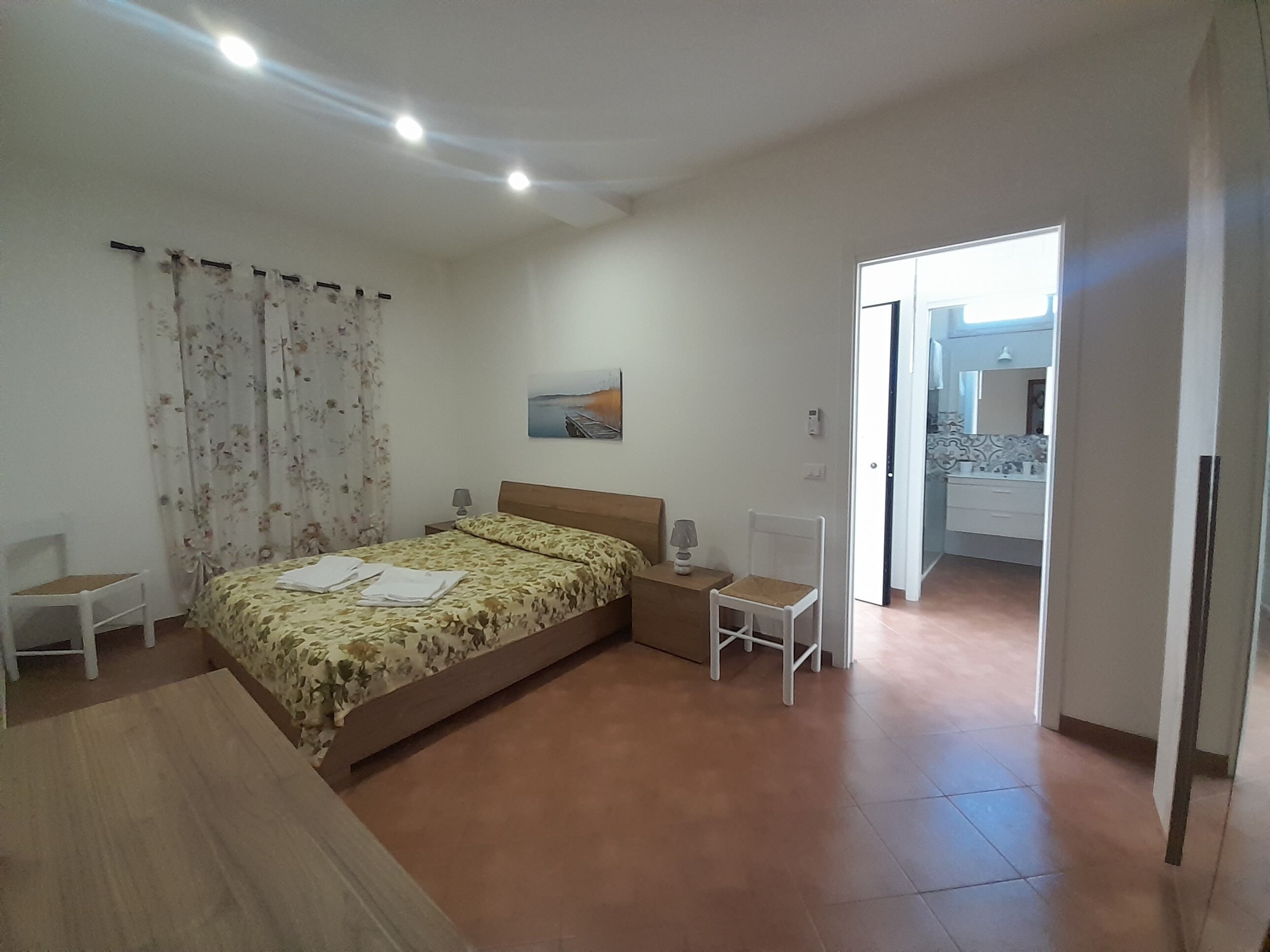 Ein Schlafzimmer mit Bett, Sessel, Schreibtisch, einem Bild und einem durchgegangenen Tür sichtbaren Badezimmer.