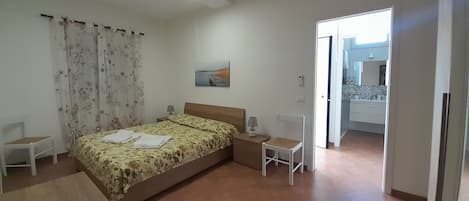 A bedroom with a bed, a chair, a desk, a painting, and a bathroom visible through an open door.