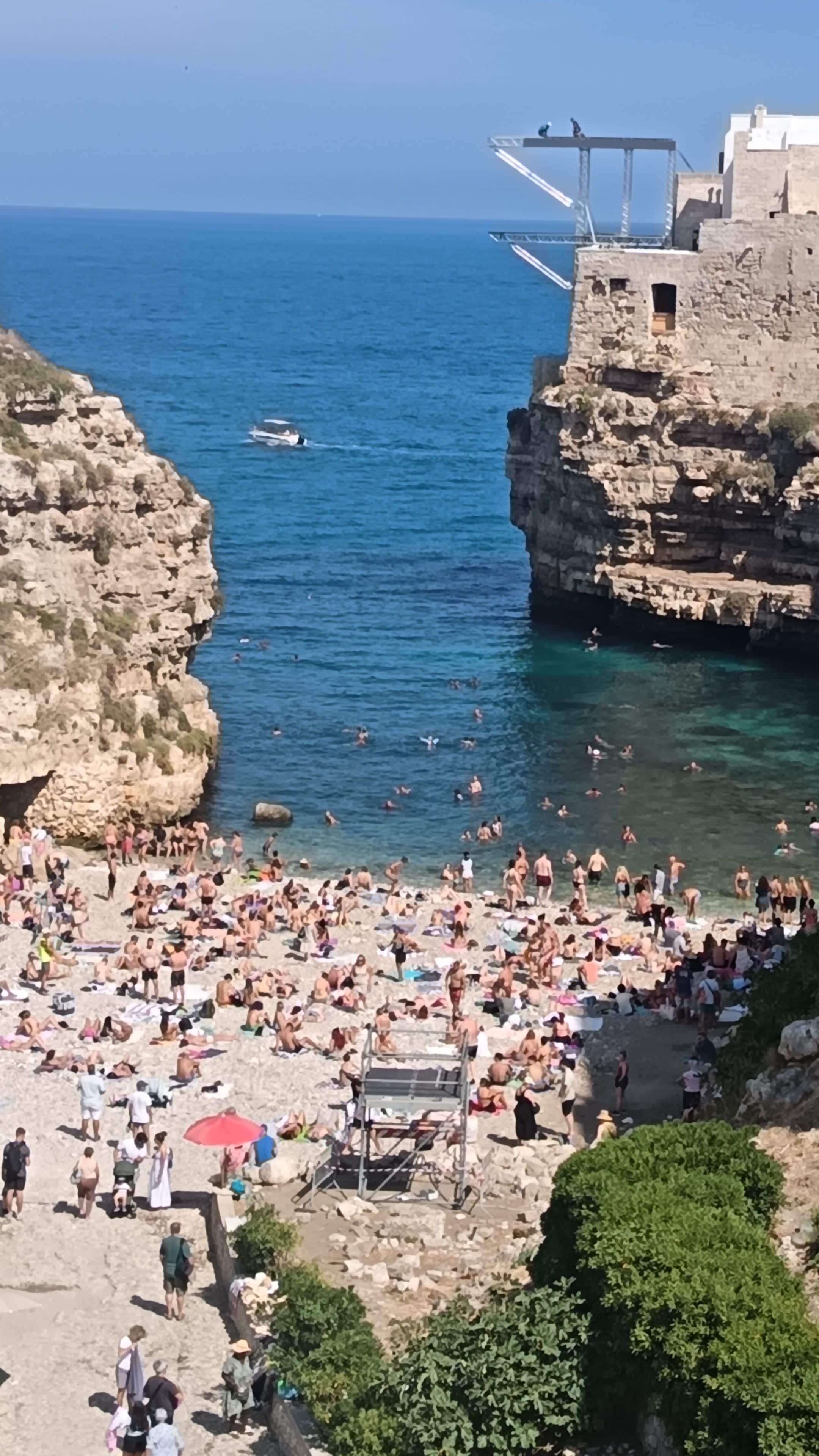 A crowded beach with people sunbathing and swimming, a rocky cliff, and a bridge structure.