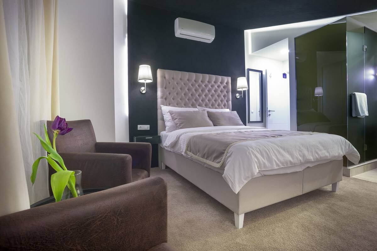 Une chambre d’hôtel moderne dotée d’un grand lit, d’un coin salon avec un fauteuil, d’une cloison en verre et d’une petite table sur laquelle est posé un vase de fleurs.