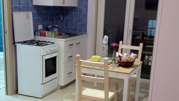 A small, well-lit kitchen with white cabinets, a stove, and a sink. There is a dining table with chairs and a window in the background. - Lilla'S House Apartment (Caltanissetta)