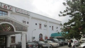 A white building with a sign that reads "ACE HOTEL". There are several cars parked in front and a person sitting on a bench. - Crystal Palace Hotel (Abuja)