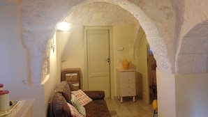 A cozy living room with a stone archway, a patterned sofa, a small wooden cabinet, and a door leading to another room.