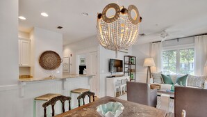 A dining area with a wooden table, chairs, and a decorative bowl. There is a chandelier above the table, and a kitchen area is visible in the background.