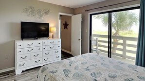 A bedroom with a bed, a TV on a dresser, and a balcony with a palm tree. - Coastal Oasis w/ Amazing Views Just Steps From The Sand + Pool Access (Tybee Island)