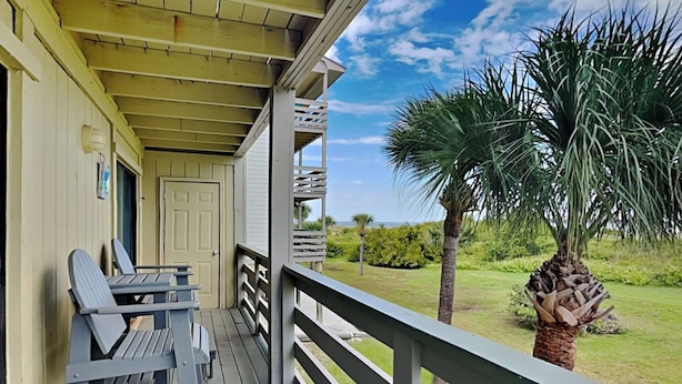 A wooden porch with two chairs and a table, overlooking a grassy area with palm trees and a clear sky. - Coastal Oasis w/ Amazing Views Just Steps From The Sand + Pool Access (Tybee Island)