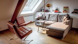 Un salon de style industriel, avec un plafond haut, un plafond en pente et une lucarne. On y trouve un fauteuil en cuir marron, un canapé gris et une petite table ronde sur laquelle est posé un vase de fleurs.