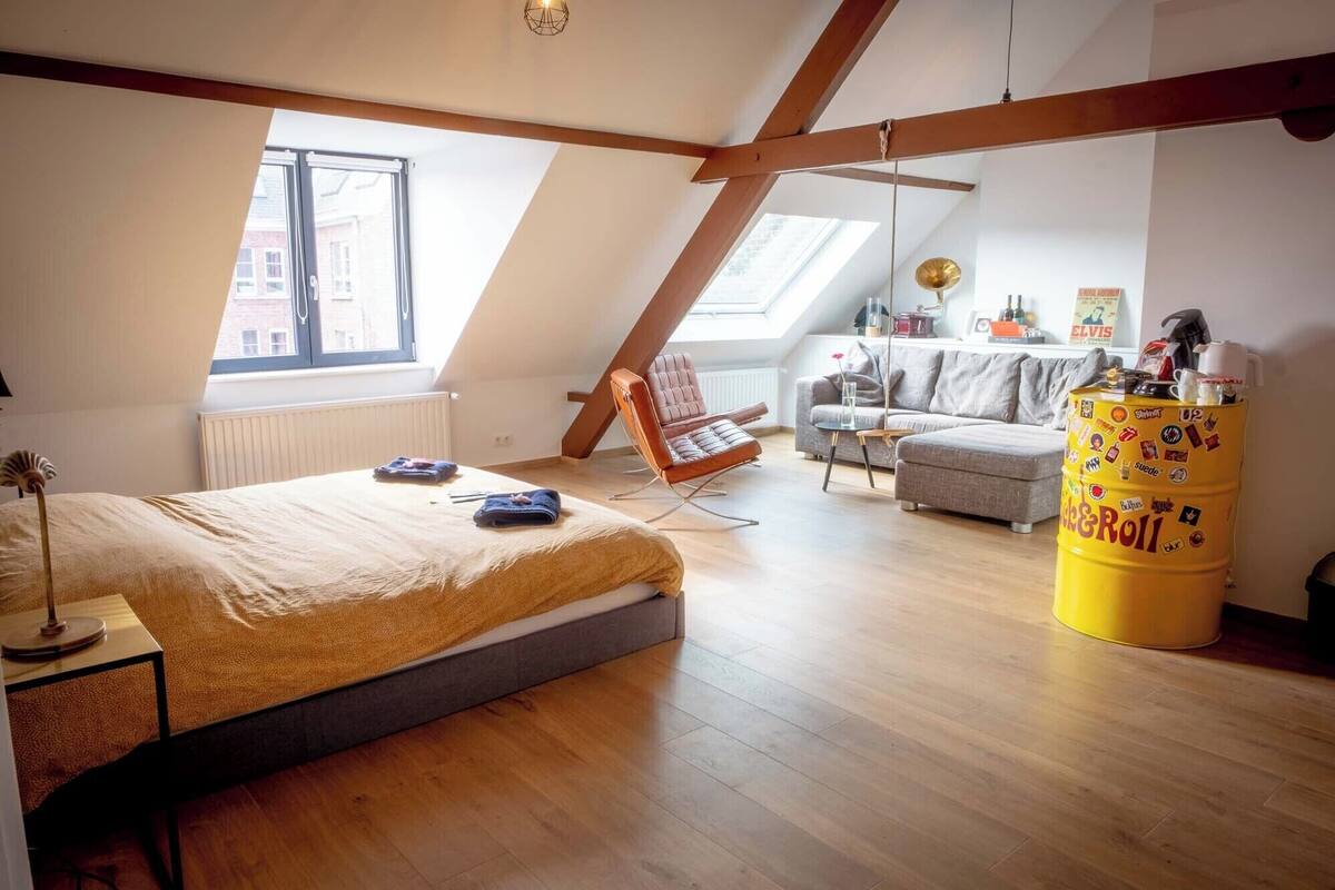 A loft bedroom with a sloped ceiling, a bed, a chair, a sofa, and a yellow barrel.