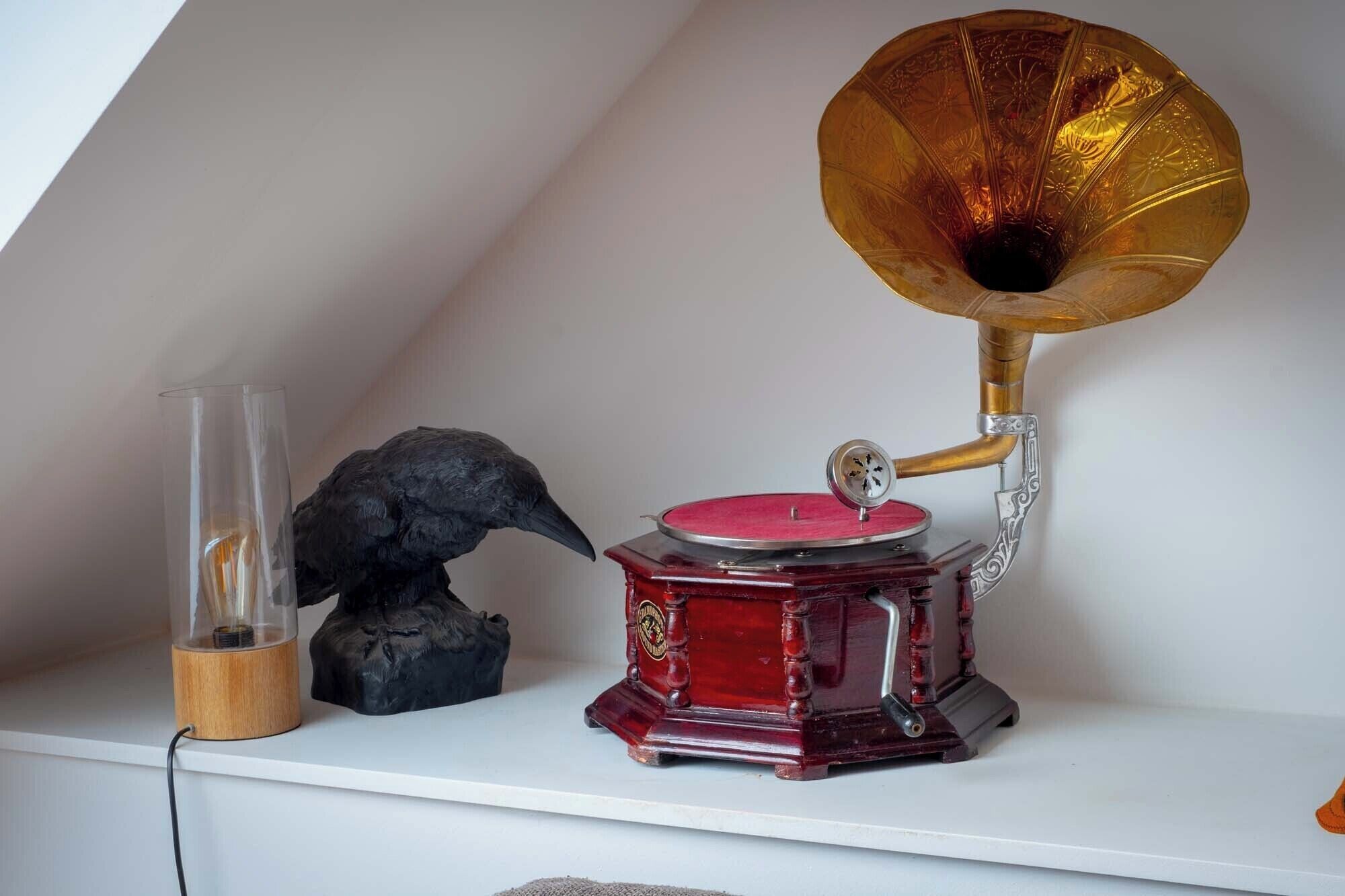Un gramófono antiguo sobre una estantería blanca, un florero de vidrio con una escultura de un pájaro y un objeto de madera.