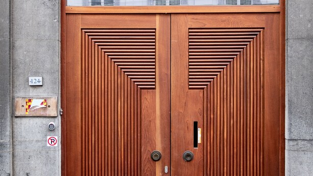Une porte en bois à deux battants ornée d’un motif géométrique, une boîte aux lettres et un panneau indiquant qu’il est interdit de se garer.