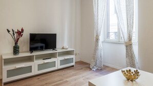 Un soggiorno moderno con una TV a schermo piatto su un mobile bianco, un vaso con piante essiccate e una finestra con tende a motivi.