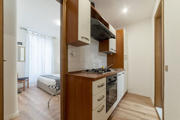 A compact kitchen with a stove, oven, and white cabinets. A dining area with a chair and a small table is visible in the background.