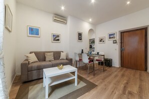 A living room with a sofa, a dining table, and chairs. There is a white coffee table with a decorative bowl. The room has white walls, wooden flooring, and an air conditioning unit.