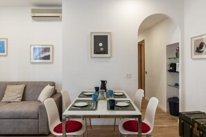 A dining area with a table set for four, white chairs with red cushions, a grey sofa, and framed artwork on the walls.