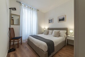 Un dormitorio con cama, lámparas de noche, una silla, un escritorio, un espejo y dos cuadros en la pared.