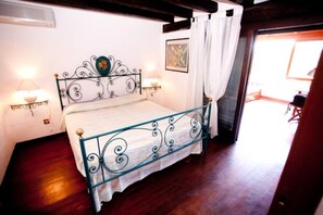 A bedroom with a metal-framed bed, white curtains, a painting on the wall, and a wooden floor. - Two-room Penthouse Pelican (Ansedonia Orbetello, Grosseto)