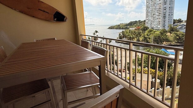A balcony with a table and chairs, a view of a tall building, and a body of water.