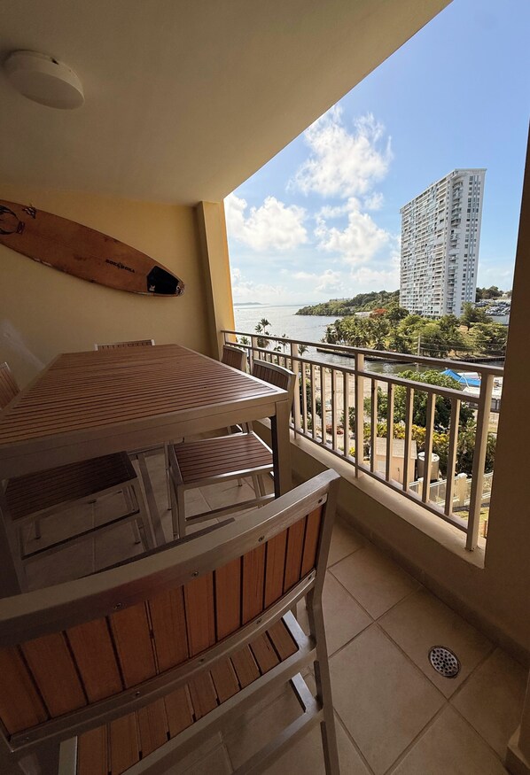 A balcony with a table and chairs, a view of a tall building, and a body of water.