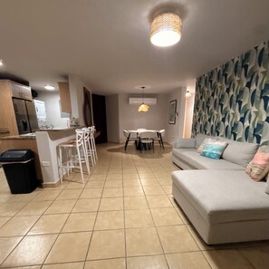 Un salon avec un canapé, une table à manger avec des chaises, un coin cuisine avec des placards et une poubelle.