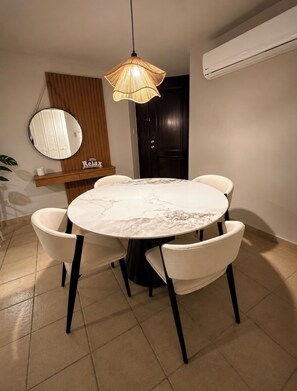 Un espace repas comprenant une table ronde en marbre, quatre chaises, un luminaire suspendu, un miroir et un appareil de climatisation.