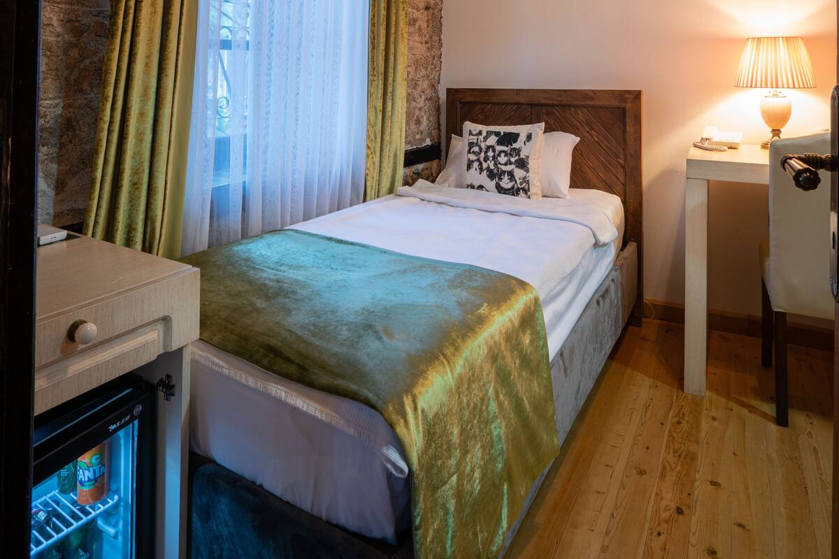 A bedroom with a bed, a bedside table with a lamp, a small refrigerator, and a window with curtains.