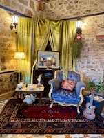 Un cuarto acogedor con pared de piedra, un gran ventanal con cortinas verdes, un cuadro enmarcado, un sillón mullido, una mesita con una lámpara, un gato y una guitarra.