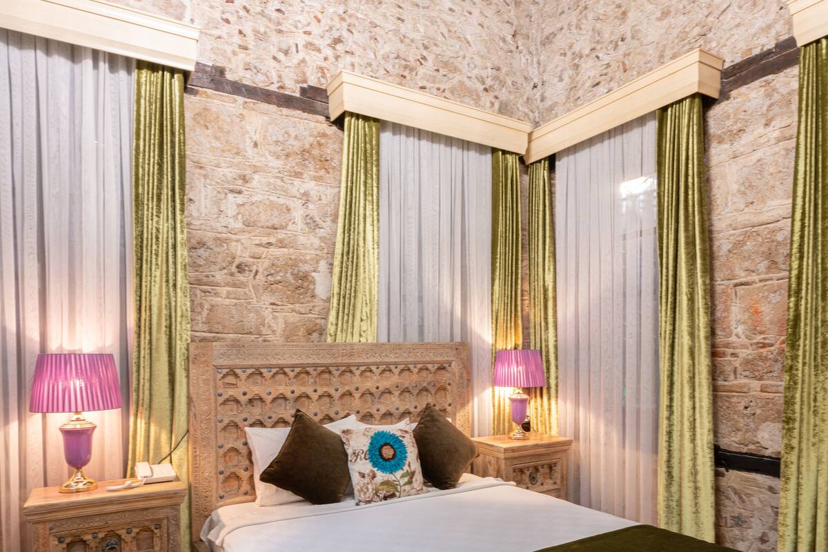 A bedroom with a stone wall, a wooden bed, green curtains, and two bedside lamps.