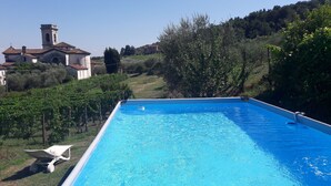 Une piscine rectangulaire à l’eau d’un bleu limpide, entourée de verdure et bordée d’une colline où se dressent des bâtiments en arrière-plan.