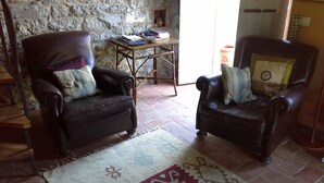 Deux fauteuils en cuir avec des coussins, une petite table en bois avec des livres et un vase, et un tapis à motifs sur un sol carrelé, avec un mur en pierre en arrière-plan.