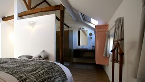 Ein Schlafzimmer mit einem großen Bett, einer Holzbalkendecke, einem Badezimmer mit Spiegel und Waschbecken sowie einer Holzlade.
