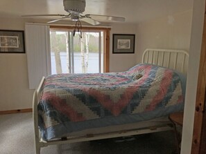 Un dormitorio con cama, ventilador de techo, ventana con persianas y dos cuadros en la pared.