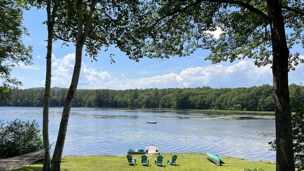 A lakeside view with a grassy area, trees, and a row of chairs and a canoe. - Enjoy Sunset views on tranquil Dexter Pond (Winthrop)