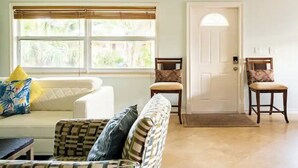 Una sala de estar con un sofá blanco, un sillón estampado y dos sillas de madera. Se ve una puerta con felpudo al fondo.