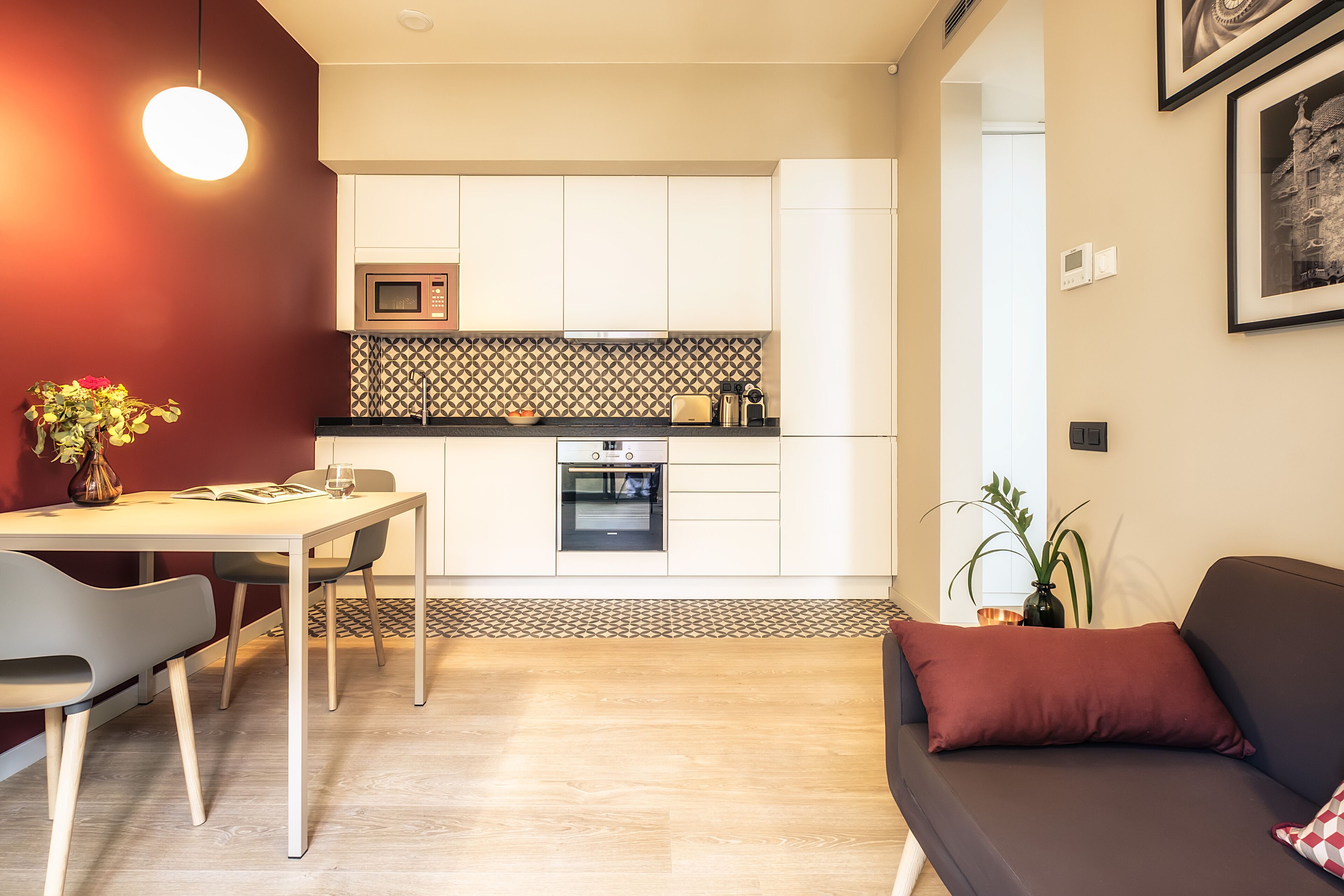 Een moderne keuken met witte kasten, een magnetron en een inbouwoven. Er is een eettafel met stoelen en een bank met een rode kussens.