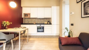 A modern kitchen with white cabinets, a microwave, and a built-in oven. There is a dining table with chairs and a sofa with a red cushion.