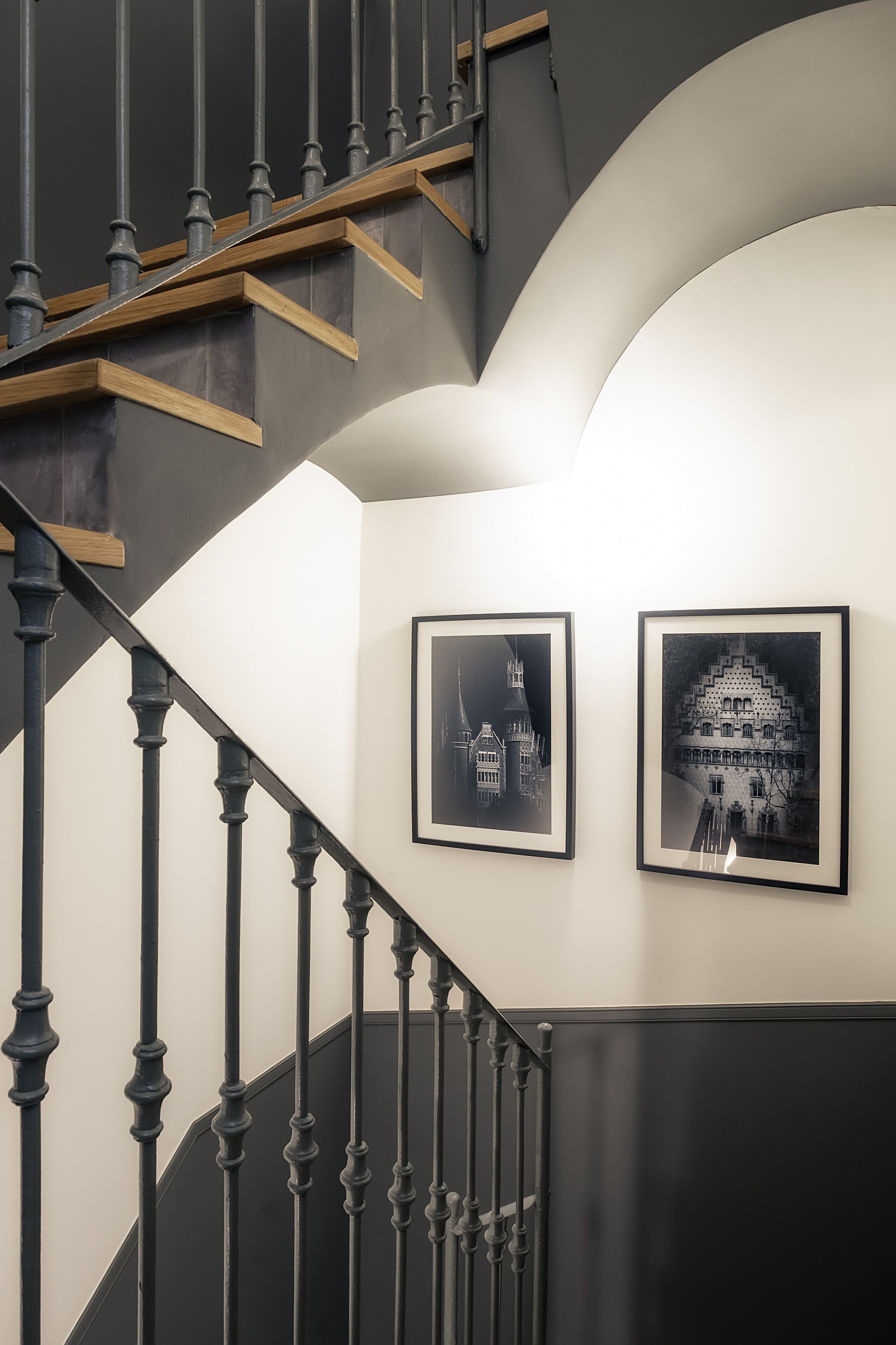 Een trap met zwarte spijlen en houten treden, die uitkomt op een plateau met twee ingelijste zwart-witfoto's aan de muur.